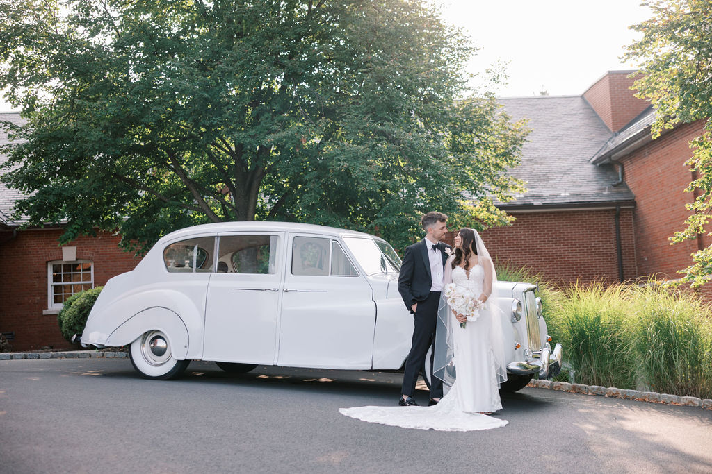a married couple in a wedding gown and tuxedo in front of a white antique car outside a brick building with a green tree